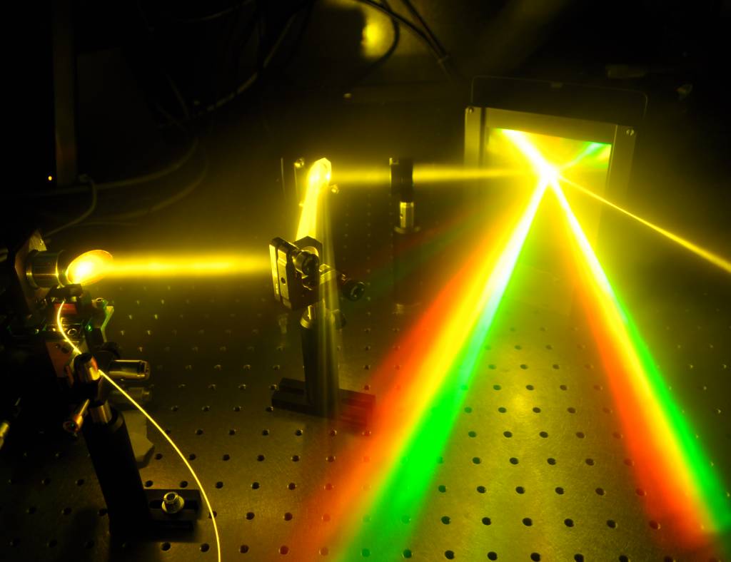 Illustration of directionality and wide spectral bandwidth of a supercontinuum source. Supercontinuum light is guided from an optical fiber into a parabolic mirror, which collimates the light. Radiation beam is white and has a diameter of 2 cm. Supercontinuum light propagates few tens of centimeters and reflects from two mirrors. Light is then focused into a diffraction grating, which reflects the wavelenghts of the supercontinuum into different directions showing two rainbows.