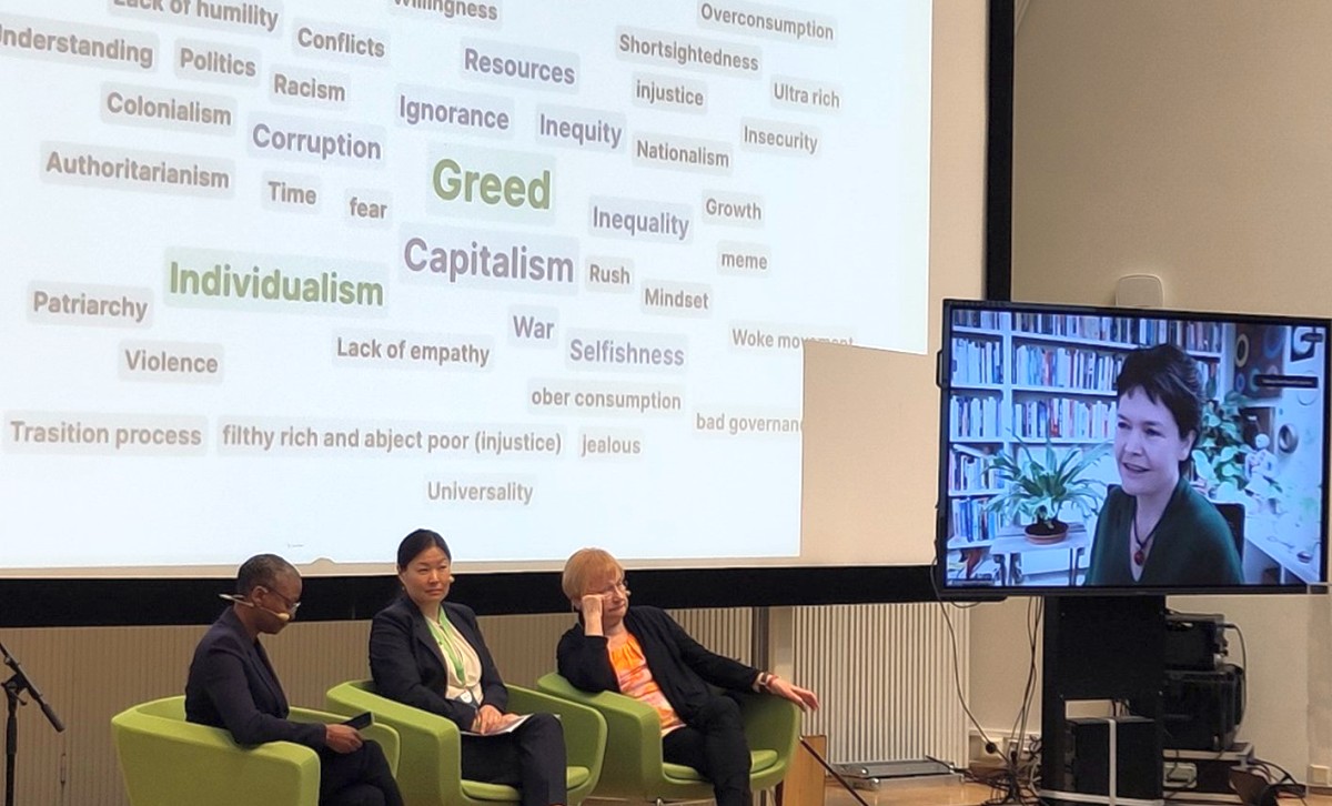 Picture from the opening plenary of SRI+SSD Congress 2024. Three speakers are sitting in chairs on a stage and one is participating remotely on a screen. Behind the speakers, a bigger screen contains a word cloud including words such as Greed, Capitalism, Individualism, Corruption, and Inequality.