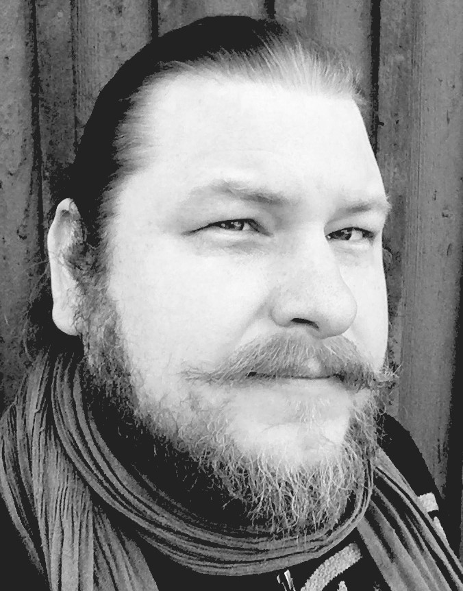 Parrakas mies katsoo suoraan kameraan mustavalkoisessa kuvassa. 