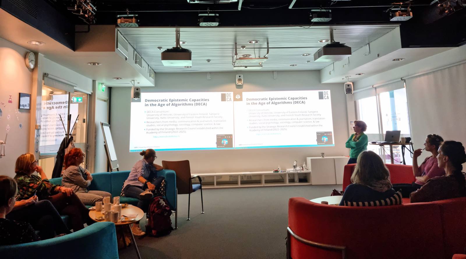 A presentation in a dim-litted space with colorful sofas. A presenter on a stage talks about a topic "Democratic Epistemic Capacities in the Age of Algorithms".