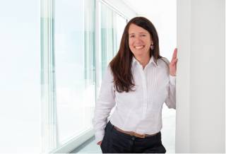 A photo of a woman with long, dark hair and a white blouse, leaning at a wall and smiling.