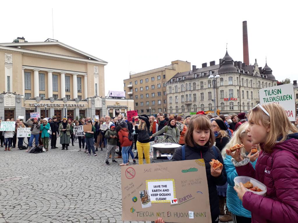 Lapsia Tampereen keskustorilla. Taustalla kaupungintalo ja etualalla lapsia kyltin kanssa, jossa lukee "Tekoja nyt, ei huomenna!".