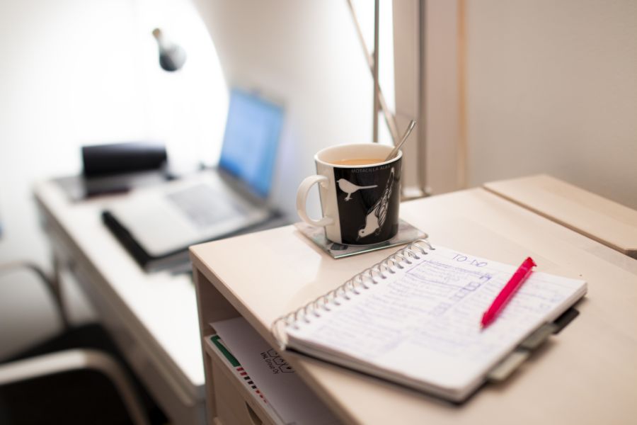 On the background open laptop. In front a coffee mug, notebook and a pen