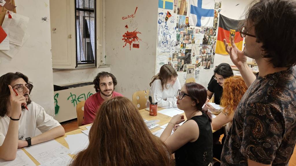 Opiskelijoita pöydän ympärillä. Kuvan etualalla näkyy takaviistosta kuvattuna Jan Pitkäsalo. Hän puhuu opiskelijoille ja elehtii samalla käsillään.