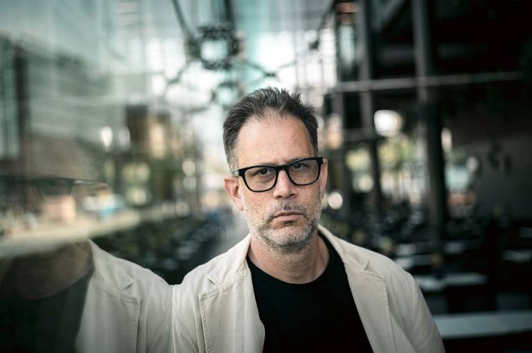 A man with glasses stands in front of a glass wall, his reflection visible.