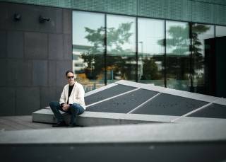 A person in sunglasses sits on a concrete structure in front of a building with large windows.