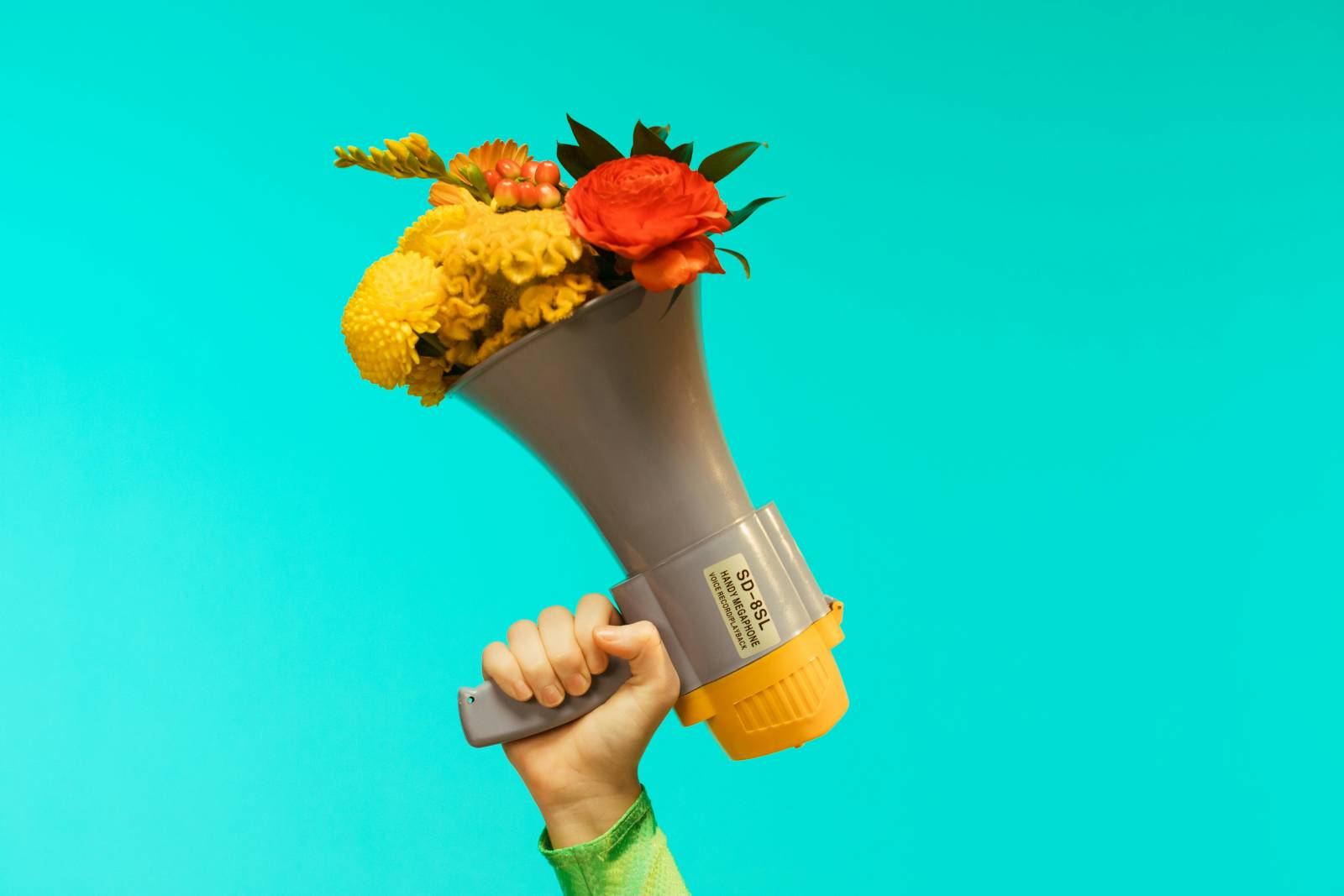 Kuvassa on turkoosia taustaa vasten kuvattu käsi, joka kannattelee ylöspäin suunnattua megafonia, jonka sisältä pursuu värikäs kukkakimppu.