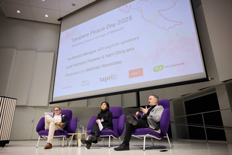 Keynote speakers and moderator sitting on stage with Peace Day poster on the background.
