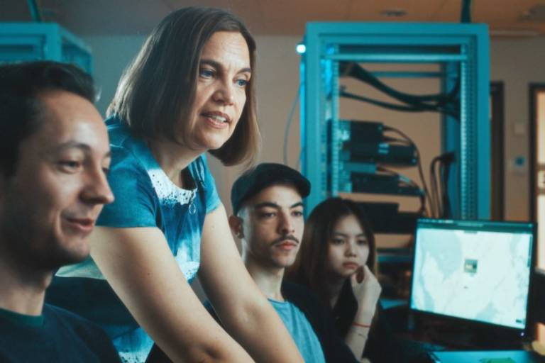 Teacher along students at a networking lab in front of monitors with racks on the background.