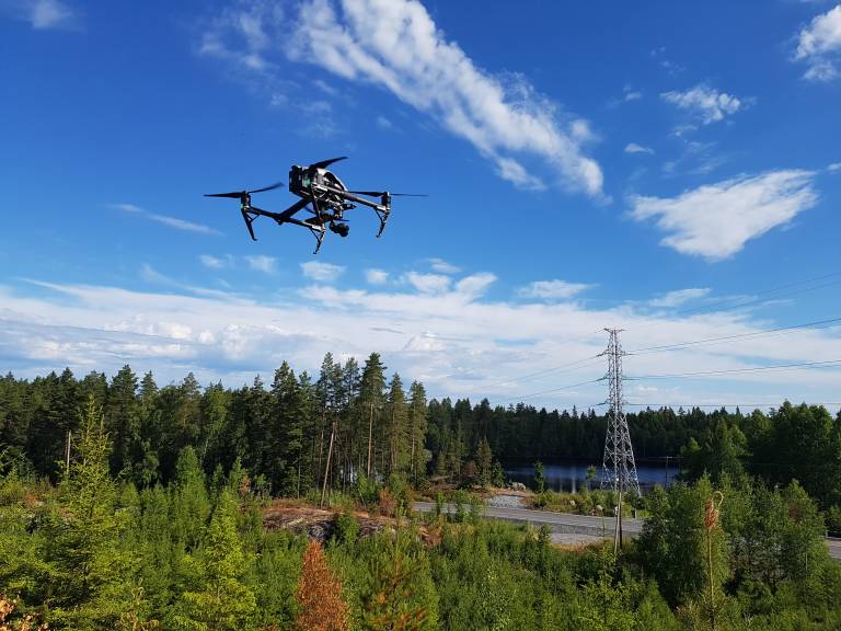 Picture of a drone in the air with sky in the background as well as power lines and trees.