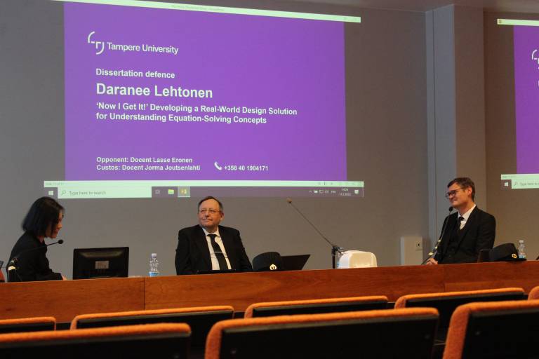 Kolme henkilöä luentosalissa, vasemmalla Daranee Lehtonen, keskellä Jorma Joutsenlahti ja oikealla Lasse Eronen. Taustalla väitöstilaisuuden diaesitys.