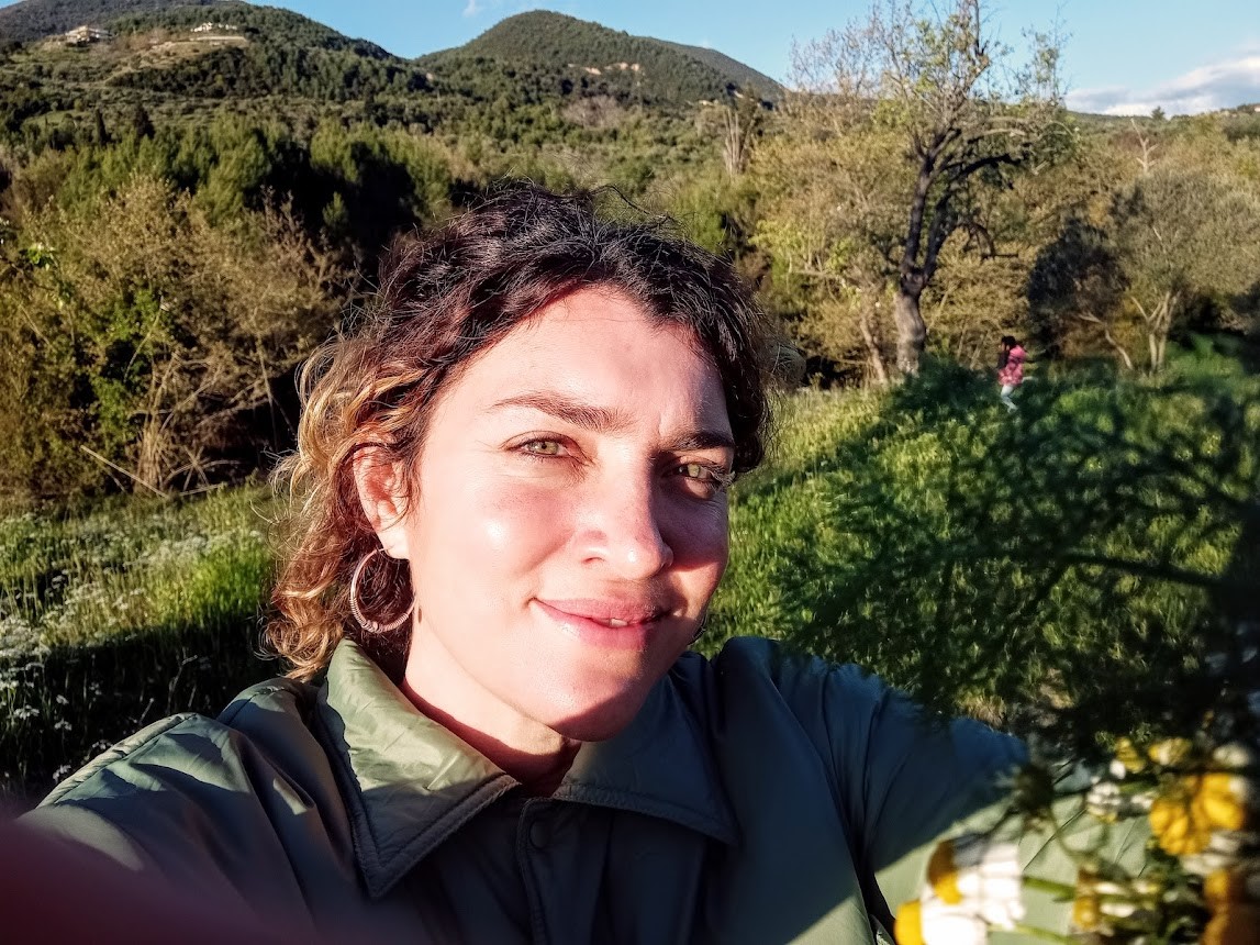 A smiling brownhaired woman. There are trees and mountains behind her.
