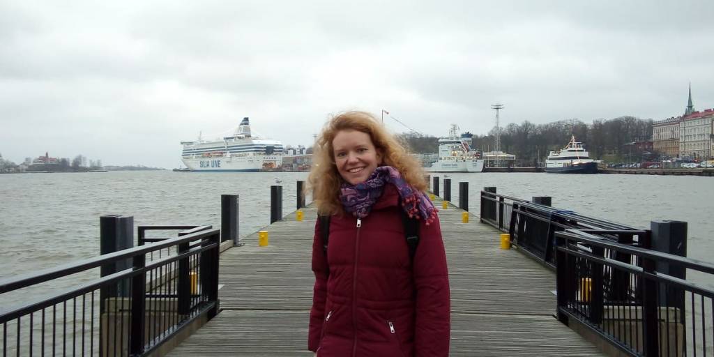 A redhaired woman standing on a pier. You can see ships and water on the background.
