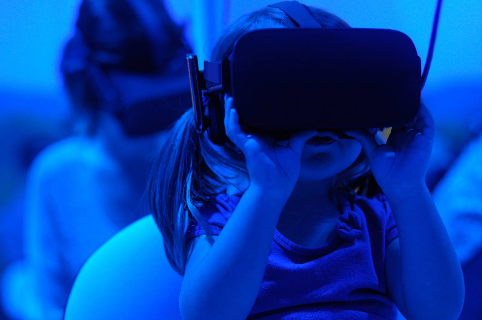 A young child with a Virtual Reality headset, clutching the mask with both their hands. Another child sits behind also wearing a Virtual Reality headset.