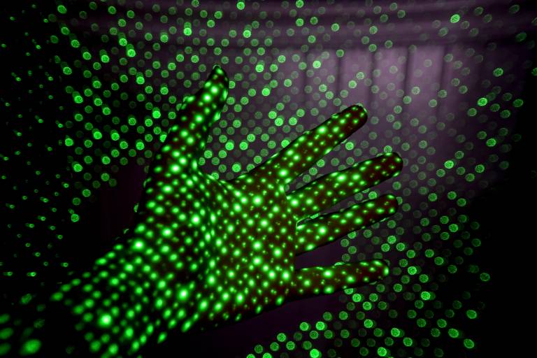 A hand covered in green dots of light, reaching out in front of a projection of light and creating a shadow on the screen behind.