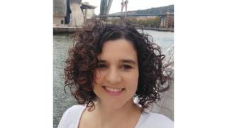 A woman with dark brown, curly hairs is smiling at the camera in a harbour scenery.