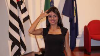 Against a white wall and wooden floor, a sign language interpreter in a black dress in the center making the sign of “person” in Brazilian sign language, with three flags behind her, one of the State of São Paulo on the left, one of Brazil in the center and one of the city of São Carlos on the right, with a red armchair next to it.