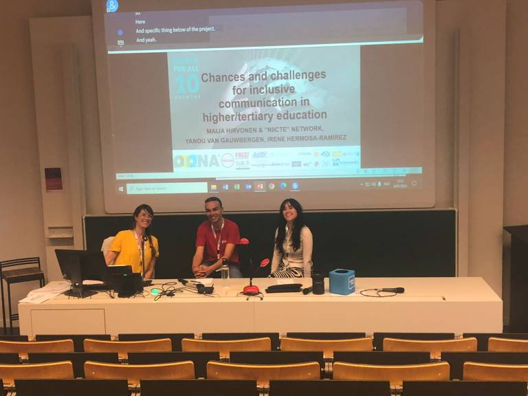 Three smiling persons behind a table in front of a lecture hall getting ready to give a presentation