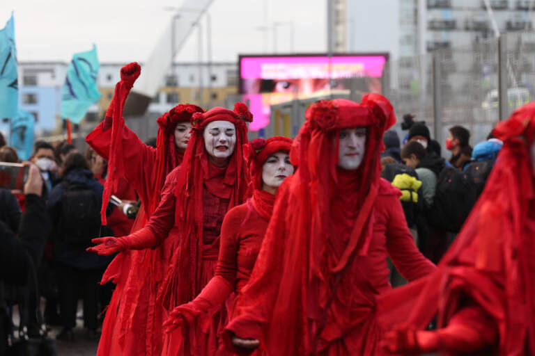 Neljä kirkkaan punaisiin vaatteisiin ja päähineisiin pukeutunutta mielenosoittajaa kulkueessa, jota ihmiset seuraavat kadun varrella.