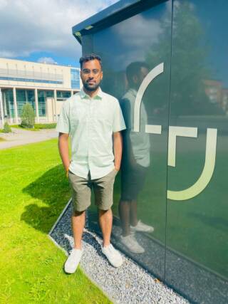 A man (sudharsana) standing near a wall with Tampere University logo, green grass background.