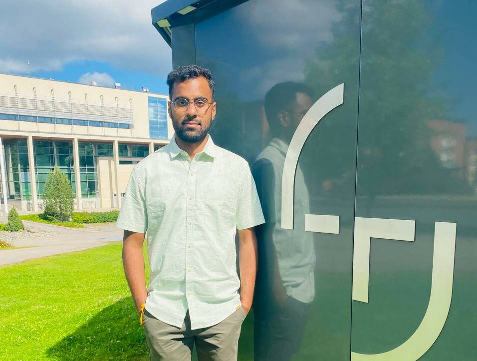 A man (sudharsana) standing near a wall with Tampere University logo, green grass background.